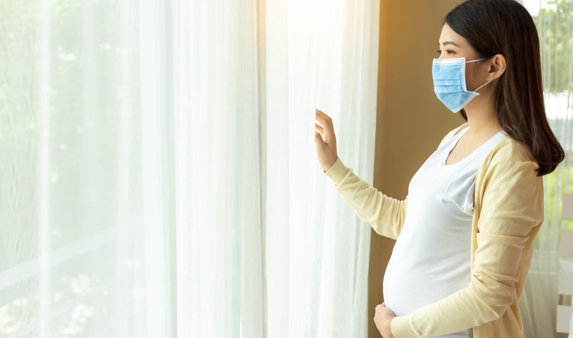 Pregnant Asian woman with mask looking out of window